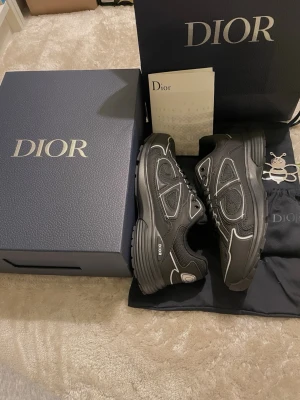 Svarta Dior B30 sneakers - Säljer ett par svarta Dior B30 sneakers med vita detaljer och reflekterande inslag. Skorna har en chunky sula, meshpaneler och klassisk snörning. Märkets logga syns tydligt på sidan och plösen. Perfekta för dig som vill ha en exklusiv och trendig look.