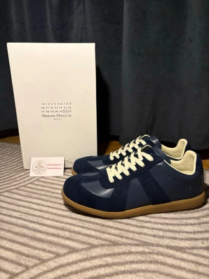 Blå Maison Margiela sneakers - Snygga sneakers från Maison Margiela i mörkblå mocka och läder med vita snören och beige innersula. Klassisk siluett med gummisula som ger en retrovibe. Perfekta för dig som vill ha en stilren och trendig look.