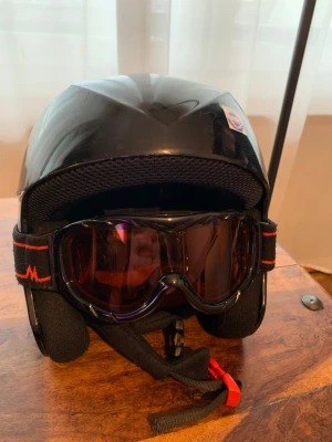 Svart skid- och snowboardhjälm med goggles - Säljer en svart hjälm för skidåkning eller snowboard med tillhörande goggles. Hjälmen har ventilationshål på sidorna och justerbart hakband. Gogglesen har svart ram och röd detalj på bandet. Perfekt för dig som vill ha både skydd och stil i backen.