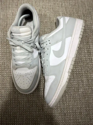 Nike Dunk Low vita och grå sneakers - Nike Dunk Low sneakers i vitt och ljusgrått med klassisk Swoosh på sidan. Ovandelen är i läder med perforerad tåbox och matchande vita och grå skosnören. Yttersulan är vit med grå detaljer. Perfekt för dig som gillar stilrena och tidlösa sneakers.