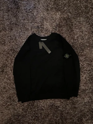 Svart sweatshirt från Stone Island - Snygg svart sweatshirt från Stone Island med klassisk rund hals och ikonisk logopatch på vänster ärm. Tröjan har ribbade muddar vid ärmslut och nederkant. Perfekt för en stilren och avslappnad look. Förpackning finns med!