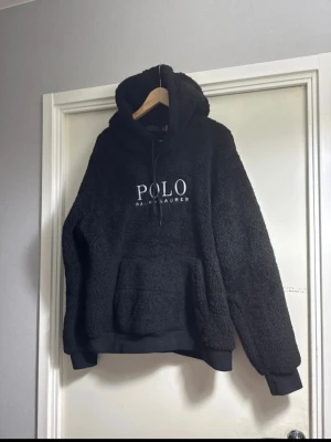 Svart teddyhoodie från Polo Ralph Lauren - Mysig svart hoodie i teddyfleece från Polo Ralph Lauren med stor huva och vit logga broderad på bröstet. Hoodien har känguruficka framtill och ribbade muddar vid ärmslut och nederkant. Perfekt för dig som gillar en avslappnad och trendig stil.