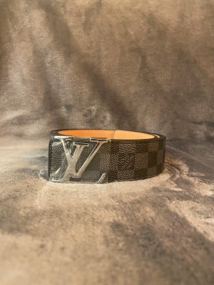 Svart rutigt LV-bälte i skinn - Snyggt svart bälte från Louis Vuitton med klassiskt rutigt mönster och stor silverfärgad LV-logga som spänne. Bältet är gjort i skinn och har en stilren design som passar till många olika outfits.