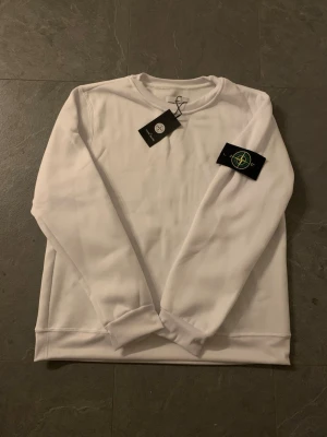 Vit sweatshirt från Stone Island - Snygg vit sweatshirt från Stone Island med klassisk rund hals och ribbade muddar. Tröjan har den ikoniska Stone Island-loggan på vänster ärm och är tillverkad i mjukt material för en clean och stilren look.