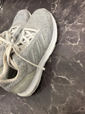 Gråa Adidas sneakers med snörning - Snygga sneakers från Adidas i ljusgrått med stickad ovandel och klassiska tre ränder på sidan. Skorna har vit sula och snörning framtill. Perfekta för en sportig och avslappnad stil.