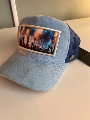 Blå Maggiore keps - Snygg ljusblå keps från Maggiore med mörkblå mesh baktill och justerbar snapback. Framtill finns ett färgstarkt motiv av en scen med artister och publik. Kepsen har broderad logga och är tillverkad i mjukt tyg och mesh för bra ventilation.