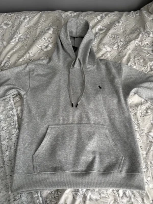 Grå hoodie från Polo Ralph Lauren - Snygg grå hoodie från Polo Ralph Lauren med klassisk huva och dragsnören. Tröjan har en stor magficka och den ikoniska broderade loggan på bröstet. Tillverkad i mjukt bomullsmaterial som känns skönt mot huden. Perfekt för en avslappnad och stilren look.