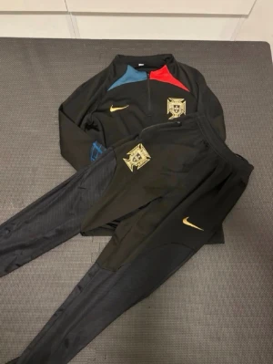 Portugal Nike träningsset svart - Snyggt svart träningsset från Nike med Portugals landslagsemblem. Tröjan har dragkedja, guldiga Nike-loggor, samt detaljer i blått och rött vid axlarna. Byxorna har smal passform och guldiga detaljer. Perfekt för fotbollsträning eller chill.