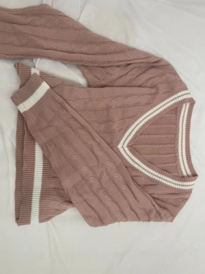 Rosa kabelstickad tröja med V-ringning - Säljer en kabelstickad tröja i dammigt rosa med vita ribbade detaljer vid ärmslut, nederkant och V-ringad hals. Tröjan har lång ärm och ett klassiskt mönster som ger en snygg retrovibe. Perfekt för dig som gillar en avslappnad men ändå stilren look.