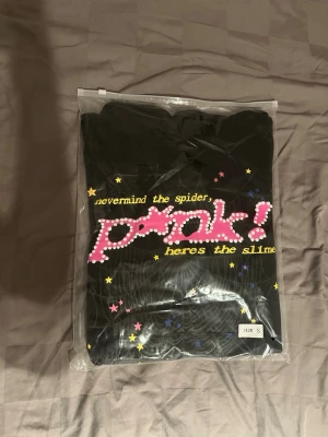 Svart Sp5der hoodie med färgglatt tryck - Svart hoodie med huva och ett stort färgglatt tryck på framsidan. Texten 'nevermind the spider, pink! heres the slime' i gult och rosa, samt stjärnor och spindelnät som detaljer. Perfekt för dig som gillar streetwear och unika prints.