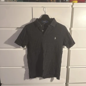 Mörkgrå piké från Polo Ralph Lauren - Mörkgrå pikétröja från Polo Ralph Lauren i slim fit med klassisk krage och två vita knappar framtill. Ikoniska broderade loggan på bröstet. Mjuk bomullskvalitet och korta ärmar gör den både snygg och bekväm. Perfekt för dig som gillar stilren design.