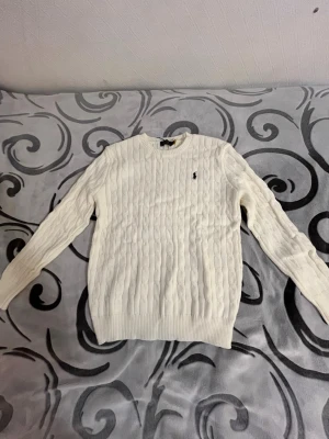 Vit kabelstickad tröja Polo Ralph Lauren - Säljer en vit kabelstickad tröja från Polo Ralph Lauren i 100% bomull. Klassisk rund halsringning, ribbade muddar och ikoniska loggan broderad på bröstet. Perfekt till lager på lager och riktigt snygg till jeans eller kjol.