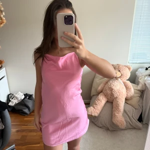 Rosa kort klänning med tunna band - Söt rosa kortklänning med tunna axelband som korsas i ryggen. Klänningen har en enkel och stilren design med lätt drapering vid halsen. Perfekt för varma dagar och somriga tillfällen.