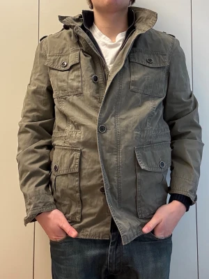 Grön vindjacka med fickor - Snygg grön vindjacka med fyra stora fickor framtill och axelklaffar. Jackan har hög krage, knappar och dragkedja framtill. Perfekt för dig som vill ha en stilren och praktisk jacka till vardags. Längd: 69cm, P2P: 52cm