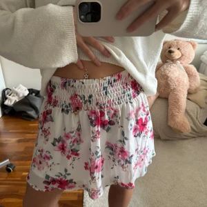 Blommig kortkjol  - Söt kortkjol i vitt med blommigt mönster i rosa. Kjolen har bred, smockad midja som ger en bekväm passform och ett flowy fall. Perfekt för varma dagar och enkel att styla med en enkel topp eller med en stickad tröja.