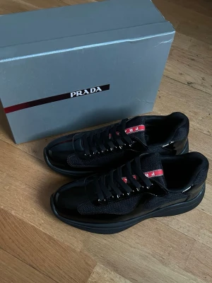 Svarta prada skor - Snygga svarta  Prada skor med glansiga och mesh-detaljer. Skorna har en tydlig röd logga på plösen och sulan, samt svarta skosnören. Klassisk låg modell som passar till streetwear och modeintresserade.