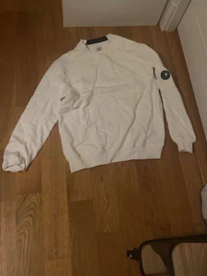 Vit sweatshirt från C.P. Company - Vit sweatshirt från C.P. Company med rund halsringning och ribbade muddar. Klassisk design med den ikoniska linsen på ena ärmen. Perfekt för en stilren och avslappnad look. Kontakta vid minsta fråga!