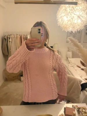 Ljusrosa kabelstickad tröja - Mysig ljusrosa stickad tröja med kabelstickade detaljer längs ärmar och sidor. Tröjan har rund halsringning och ribbstickade muddar vid ärmslut och nederkant. Perfekt för att hålla värmen under kyliga dagar.
