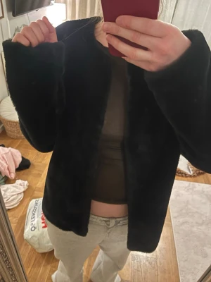 Svart fuskpälsjacka i klassisk modell - Säljer en svart fuskpälsjacka med rak passform och långa ärmar. Jackan har en stilren look utan synliga detaljer och är perfekt för dig som vill ha en enkel men trendig pälsjacka. Materialet är mjukt och ger en lyxig känsla.