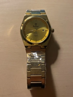 herrklocka kvartz i metall  - Stilren klocka med guldfärgad urtavla. Klassisk design med datumvisning och kvartsverk. Perfekt accessoar för dig som gillar en tidlös look med lite extra bling.