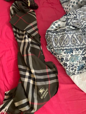 Burberry halsduk - Snygg halsduk från Burberry i mörkbrunt med klassiskt rutmönster i beige, rött och svart. Halsduken har fransar i ändarna och Burberry London-logga broderad i gult. Perfekt accessoar för att lyfta din outfit.