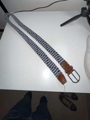 Blåvitt flätat bälte med läderdetaljer - Snyggt blåvitt flätat bälte med bruna läderdetaljer och silverfärgat spänne. Bältet har ett klassiskt mönster och passar perfekt till jeans eller chinos. Materialet är elastiskt tyg kombinerat med läder vid ändarna och spännet. Pris kan sänkas😀