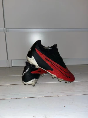 Nike Phantom GT2 Elite fotbollsskor - Säljer ett par Nike Phantom GT2 Elite fotbollsskor i svart och rött med stora svarta Swoosh-loggor på sidorna. Skorna har låg profil, snörningslös design och ovandel i syntetmaterial för bra bollkontroll. Perfekta för spel på gräsplan.