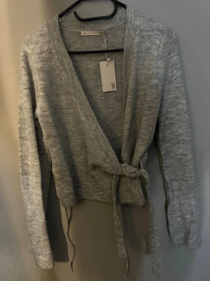 Grå omlottkofta från lager 157 - Mysig grå stickad omlottkofta från lager 157 med långa ärmar och knytband i midjan. Är alldrig använd och har lappen kvar.