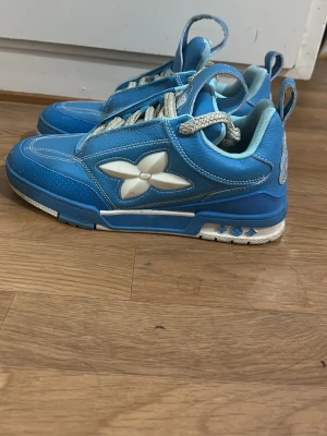 Blå Louis Vuitton sneakers - Snygga blå sneakers från Louis Vuitton med vit LV-blomma på sidan och gråa tjocka skosnören. Skorna har vita detaljer och en robust sula med blå och vita inslag. Perfekta för dig som vill sticka ut med exklusiva och trendiga sneakers.