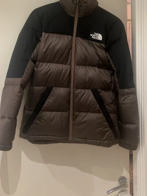 Brun dunjacka från The North Face - Snygg brun och svart dunjacka från The North Face med hög krage och dragkedja framtill. Jackan har två snedställda fickor med svarta detaljer och är perfekt för kalla vinterdagar. Klassisk boxig passform och diskret logga på bröstet.