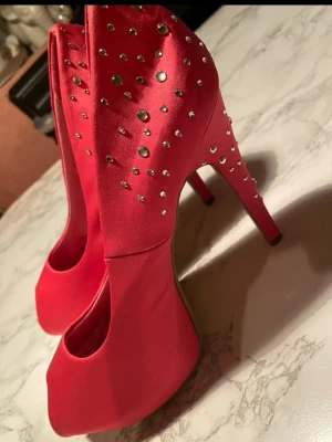 Rosa pumps med strass från Bianco - Snygga rosa pumps från Bianco med öppen tå och höga klackar. Skorna är dekorerade med glittrande stenar på hälen och sidan, vilket ger en extra glamorös känsla. Tillverkade i ett satinliknande material som känns lyxigt och trendigt.