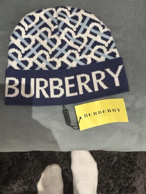 Blåvit Burberry mössa med logga - Säljer en snygg blåvit mössa från Burberry med stort logotyptryck framtill och ett mönster av sammanflätade bokstäver. Mössan är stickad i mjukt material och har en bred uppvikt kant. Perfekt accessoar för dig som gillar exklusiv streetstyle.