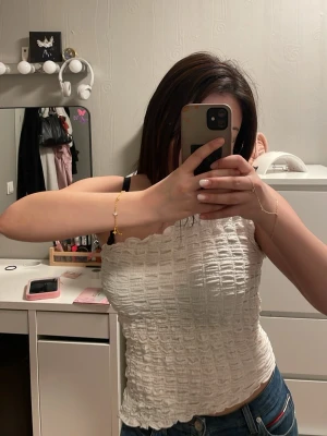 Vit oneshoulder topp i smock - Säljer en vit oneshoulder topp med smockad struktur och vågig kant upptill. Toppen är ärmlös och har en tight passform som framhäver figuren. Perfekt att styla med jeans eller kjol för en trendig look. Pris kan diskuteras