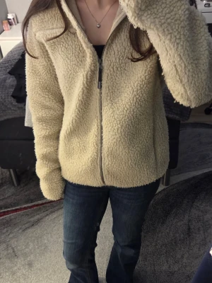 Beige teddyjacka med huva  - Mysig beige teddyjacka med huva och dragkedja framtill. Jackan har två sidofickor och är tillverkad i fluffigt teddy-material som håller dig varm under kyliga dagar. Perfekt för dig som gillar en avslappnad och trendig stil.