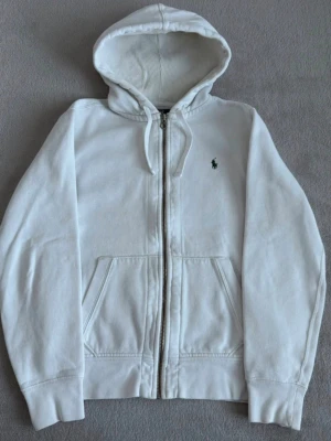 Vit hoodie från Polo Ralph Lauren - Snygg vit hoodie från Polo Ralph Lauren med dragkedja framtill och klassisk huva. Broderad logga i mörkgrönt på bröstet och två fickor fram. Tillverkad i mjuk bomull för skön känsla. Perfekt för en clean och avslappnad stil. Passar M och S