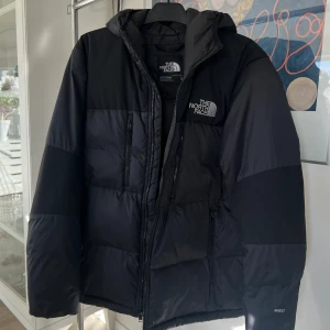 Svart dunjacka från The North Face - Säljer en svart dunjacka från The North Face med huva och dragkedja framtill. Jackan har två stora sidofickor med dragkedja och logga på både bröstet och ryggen. Lätt dunjacka som passar såväl vinter som sen höst och tidig vår. 