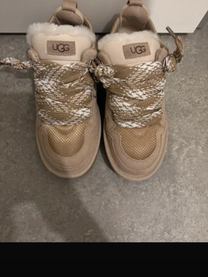 Beige sneakers från UGG med foder - Snygga beige sneakers från UGG med grova vita och beige snören, mjukt vitt foder och detaljer i mocka och mesh. Yttersulan är räfflad och i gummi, perfekt för en cool och bekväm stil. UGG-logga på plösen och hälen.