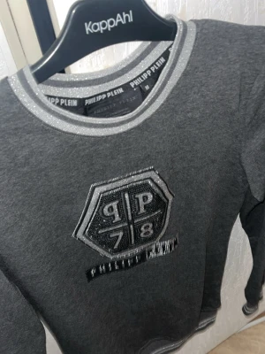 Grå sweatshirt från Philipp Plein - Snygg grå sweatshirt från Philipp Plein med silverglittriga detaljer vid krage och muddar. Stor logga och emblem med texten 'PP 78' på bröstet. Tillverkad i mjukt material, perfekt för en trendig och avslappnad look. Storlek M och köpt för 1,800kr. Som ny knappt använd. Säljer mina plagg som jag inte använder. 