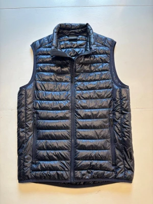 Mörkblå Everest dunväst - Stilren mörkblå dunväst med horisontella sömmar och hög krage. Västen har två fickor med dragkedja på sidorna och stängs med en hel dragkedja framtill. Perfekt för lager-på-lager och enkel att matcha till olika outfits. Storlek XS (passar även S). Skick 9/10.