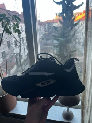 Svarta Dior B22 - Snygga svarta Dior sneakers med vita och grå detaljer. Skorna har en chunky sula och ovandel i mesh och syntetmaterial för en sportig vibe. Klassisk snörning och diskret Dior-logga på sidan. Perfekta för dig som gillar streetwear och exklusiva märken.