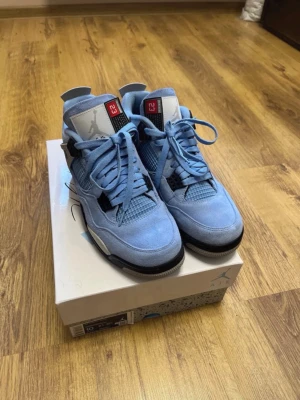 Air Jordan 4 Retro University Blue - Snygga Air Jordan 4 Retro University Blue sneakers i ljusblå mocka med svarta och grå detaljer. Klassisk siluett med meshpaneler, synliga snörhål och Jumpman-logga på plösen. Perfekta för dig som vill sticka ut med stilrena och ikoniska sneakers.