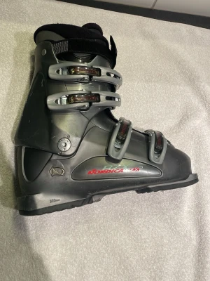 Svarta pjäxor från Nordica stl.39 - Säljer ett par svarta pjäxor från Nordica med fyra justerbara spännen med silver och röd detalj. Skalet är gjort i hårdplast och innerskon är vadderad för extra komfort. Väl fungerande men repiga.