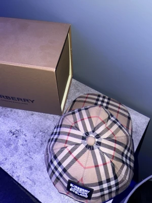 Burberry beige rutig keps - Säljer en klassisk Burberry keps i beige med det ikoniska rutmönstret i svart, vitt och rött. Kepsen har en rundad form och en liten patch framtill med Burberry London England. Tillverkad i bomull och polyester för en skön känsla.