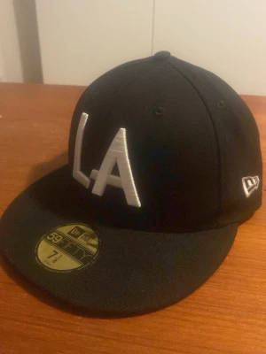 Svart LA New Era 59FIFTY keps - Snygg svart keps från New Era 59FIFTY med stor vit LA-logga framtill och ASG 2022-brodyr bak. Klassisk flat brim och broderad New Era-logga på sidan. Perfekt för dig som gillar streetwear och sportig stil.