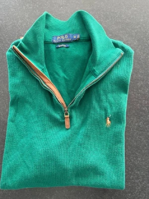 Grön quarter zip  tröja från Polo Ralph Lauren - Snygg grön tröja från Polo Ralph Lauren med quarter zip och klassisk broderad logga på bröstet. Tröjan är ribbstickad och har en hög krage med dragkedja och läderdetalj. Perfekt för en stilren och avslappnad look.