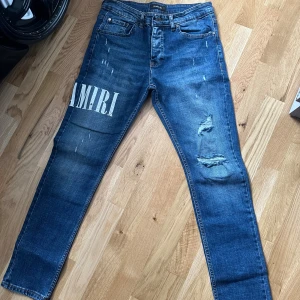 AMIRI Slim jeans med tryck - Blå skinny jeans från AMIRI med stort vitt tryck på vänster lår, slitningar och trasiga detaljer på höger ben. Klassisk femficksmodell i stretchig bomullsdenim. Snyggt tvättade med en modern, smal passform och låg midja. Inte äkta