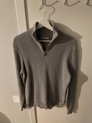 Grå stickad halvzip från Zara - Snygg grå stickad tröja från Zara med halv dragkedja och hög krage. Tröjan har ribbstickad struktur och långa ärmar, perfekt för lager-på-lager. Enkel och stilren design som passar till det mesta.