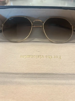 Ray-Ban guldiga solglasögon - Snygga solglasögon från Ray-Ban med tunn guldfärgad metallbåge och mörka, lätt tonade glas. Säljer pga att jag inte använder dom. Väldigt bra skick, kontakta för fler bilder