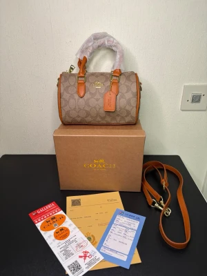 Coach beige barrel handväska - Snygg beige handväska från Coach i klassisk barrel-modell med monogrammönster och detaljer i ljusbrunt läder. Väskan har guldfärgade metalldetaljer, avtagbar axelrem och Coach-tag. Perfekt för dig som gillar stilrena accessoarer.