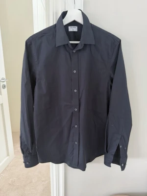 Mörkblå skjorta från Filippa K - Klassisk mörkblå skjorta från Filippa K med lång ärm och knappar framtill. Skjortan har en stilren krage och är tillverkad i ett mjukt bomullstyg som känns skönt mot huden. Perfekt för en clean och enkel look.